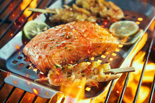 Raw salmon fillet with prawn kebabs seasoned with chili and spices on a fire grilling in a griddle over the grid in close up with slices of lemon