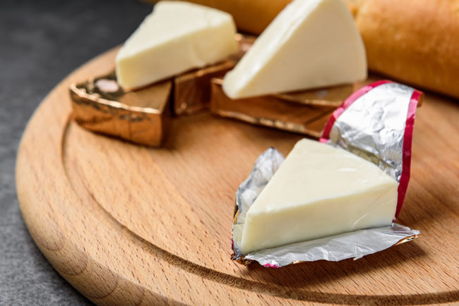 Processed triangle cheese in foil and baguette on wooden board on gray background.