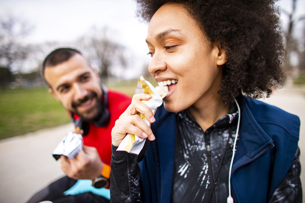 Young sportswoman enjoying in a protein bar with her partner while sitting on a bench in a city park.